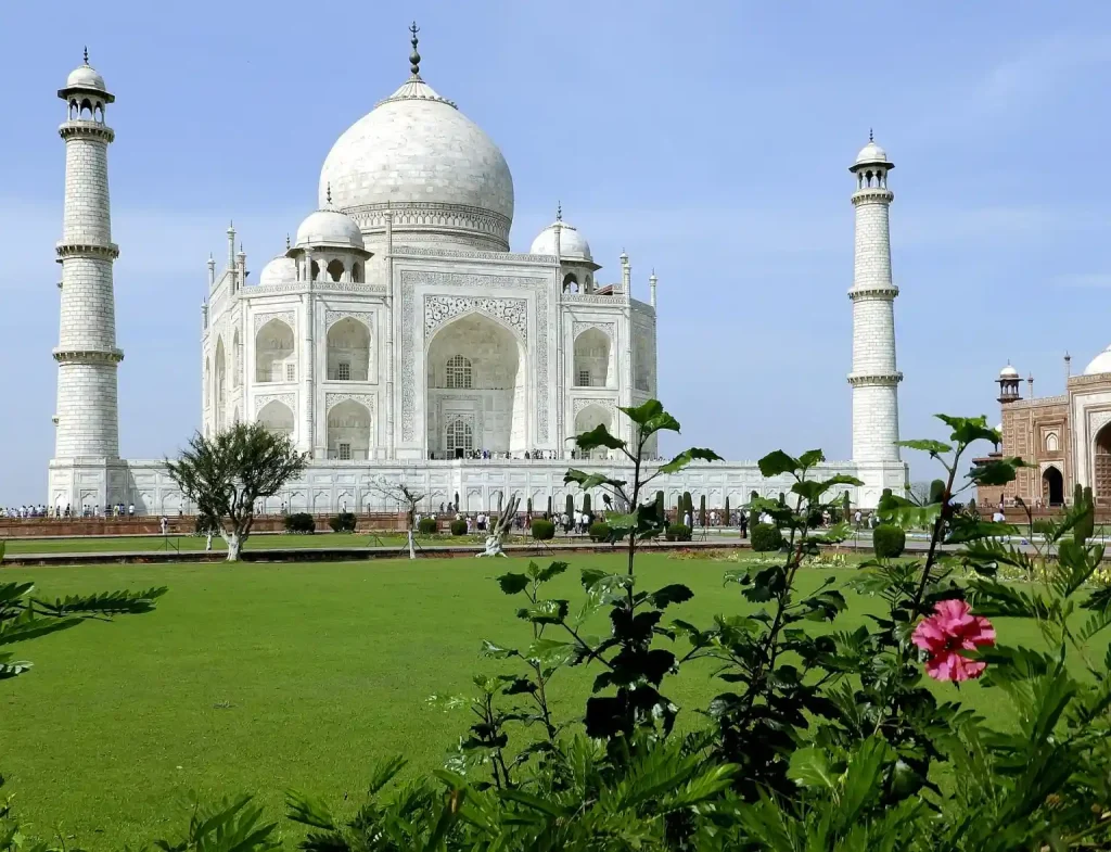 The image shows the Taj Mahal, a white marble mausoleum with a large central dome flanked by four tall minarets. Set against a clear blue sky, it stands majestically with a lush green lawn and flowering bushes in the foreground.