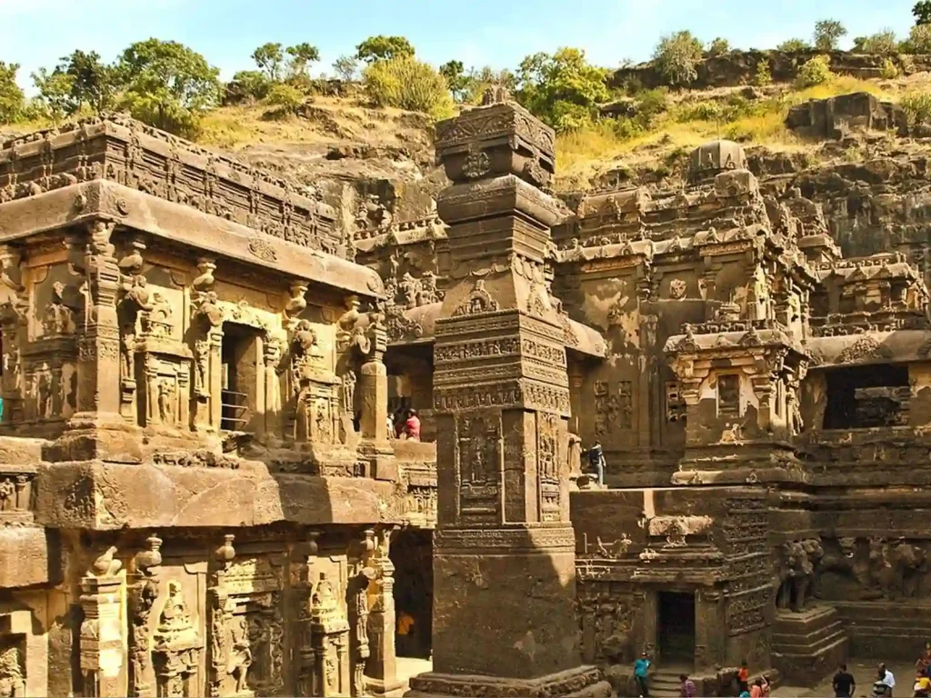 An ancient rock-cut temple complex with intricate carvings stands under a clear sky. The structures are made of stone and showcase elaborate details and pillars. People can be seen exploring the area, adding a sense of scale to the grandeur of the architecture.