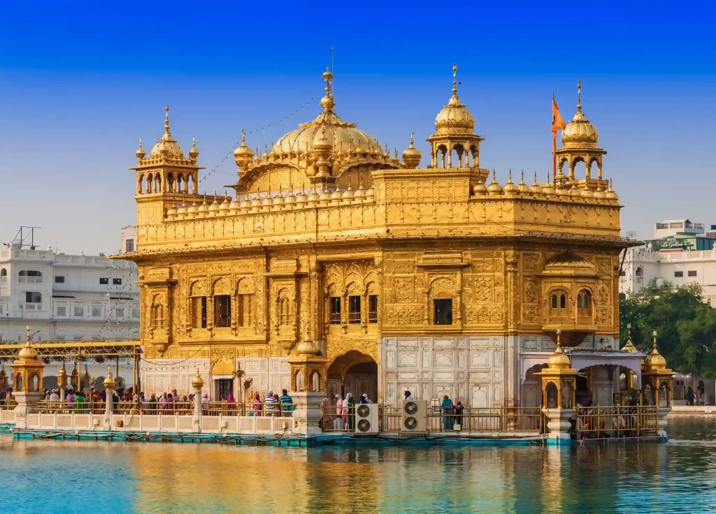 The image shows the Golden Temple in India. The temple's exterior is exquisitely covered in gold, reflecting in the surrounding water. The sky is clear and blue, with some visitors seen near the entrance and the temple’s walkways.