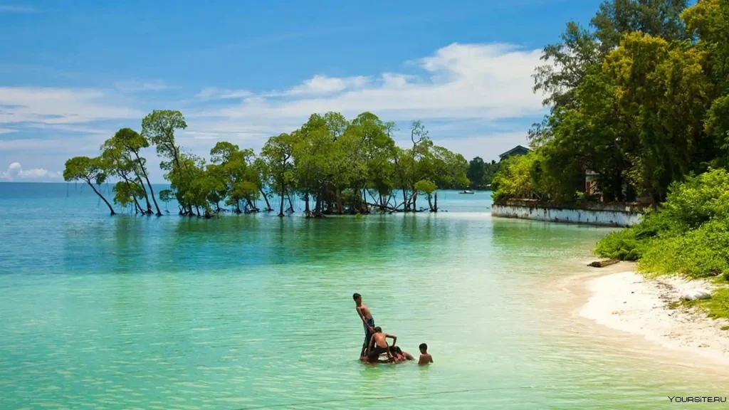 A tropical beach scene with three people enjoying the shallow, clear turquoise water. In the background, small islets with green trees are visible. A mix of sandy shorelines and lush greenery surrounds the tranquil bay. The sky is blue with scattered clouds.