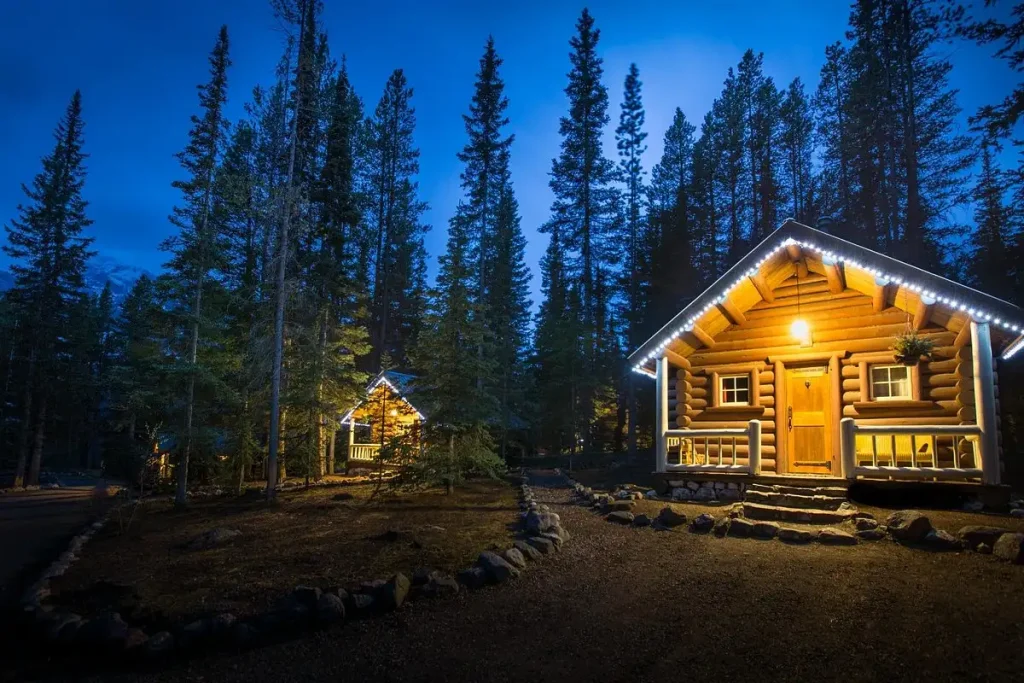 A cozy log cabin with illuminated eaves sits amid tall pine trees at dusk. Another brightly lit cabin is visible in the background. The scene exudes a serene, rustic charm with the glow of the lights contrasting against the deep blue twilight sky, creating an inviting atmosphere.