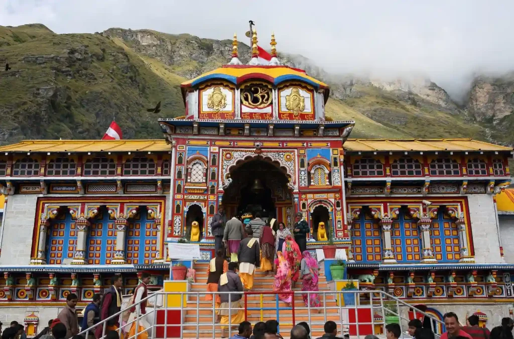 A group of people ascends the steps to the main entrance of a brightly colored, intricately decorated temple situated at the base of a mountain. The temple features vibrant patterns and statues, with lush greenery and mist-covered peaks in the background.