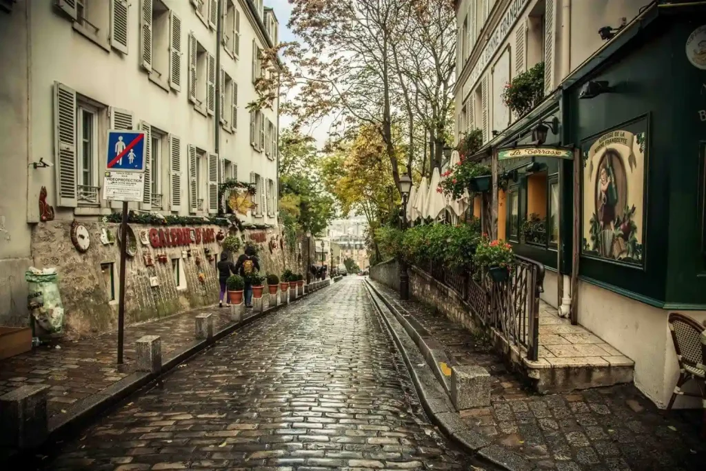 A narrow, cobblestone street in a quaint European town, reminiscent of those you'll explore on the streets of Paris, flanked by charming buildings with shutters and greenery. The gently sloping street is lined with cozy cafes, small shops, and planters. Trees with autumn leaves add a touch of color to the scene.