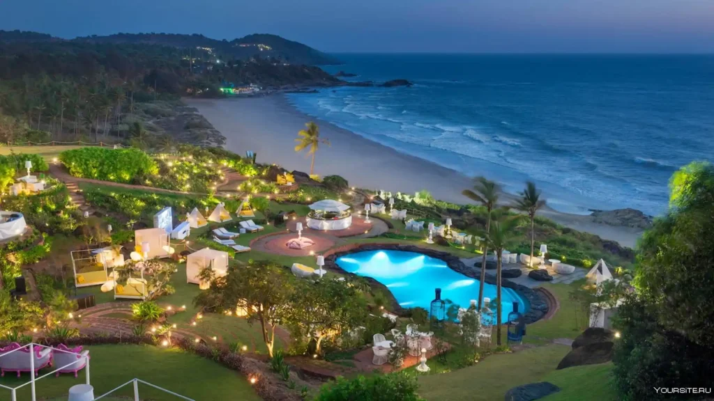 A picturesque beachfront resort at dusk, featuring landscaped gardens, illuminated pathways, cabanas, and a large, glowing blue pool. The sandy beach and rolling waves of the ocean are visible in the background, with hills and vegetation on the horizon.