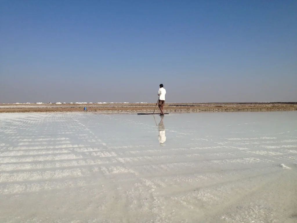 A person in a white shirt and shorts is walking on a reflective surface with a tool in their hand. The sky is clear and blue, and the horizon is visible in the background.