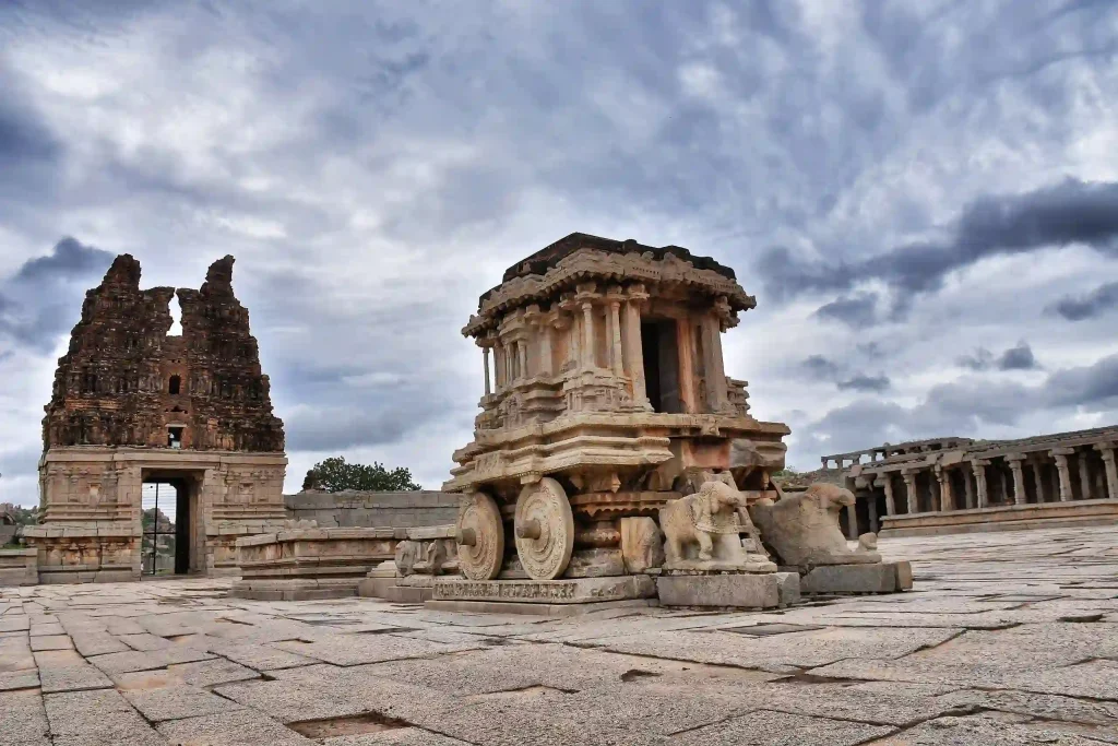 A monumental stone chariot stands prominently in a courtyard with ancient ruins and columns under a cloudy sky. The intricately carved chariot, made of granite, features wheels and detailed sculptures, exemplifying rich historical architecture.