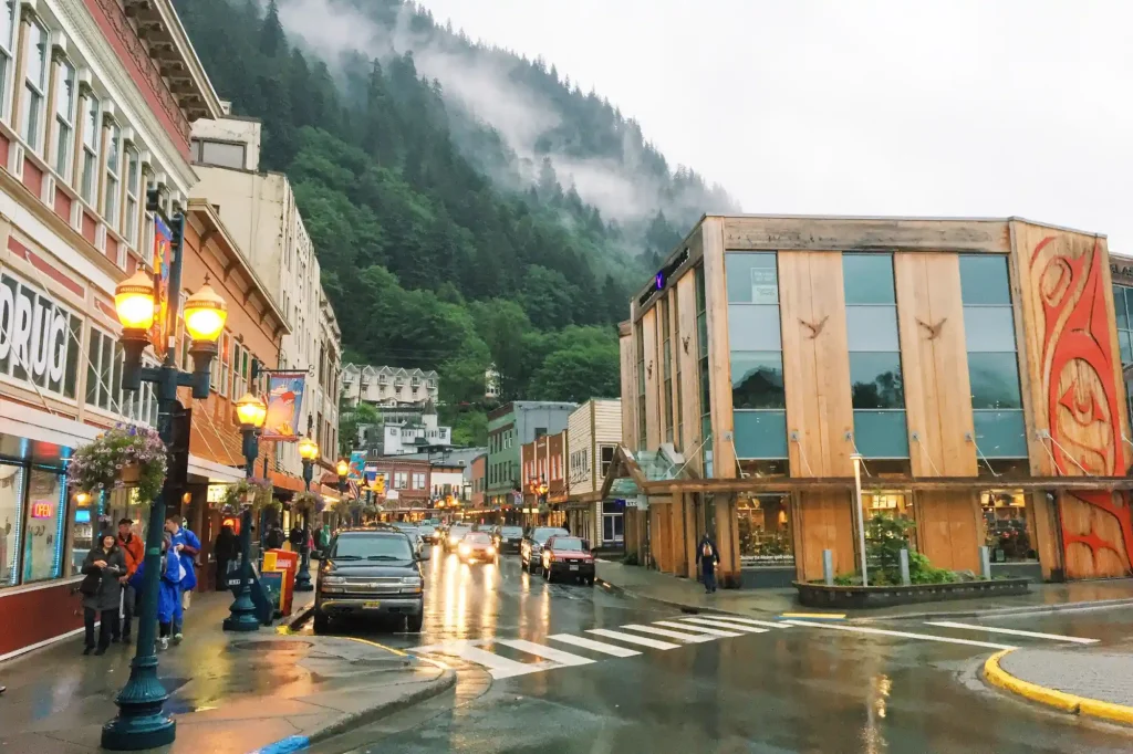 A rainy street in Juneau, Alaska, features colorful buildings. People with umbrellas walk on sidewalks, while cars drive on wet roads. A mist-covered forested hill rises in the background, and streetlights illuminate the scene.