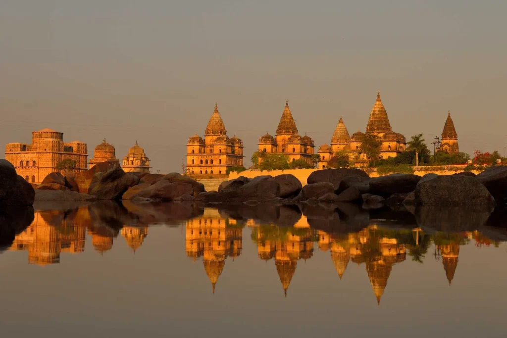 A tranquil scene of ancient temples bathed in golden sunlight, reflecting perfectly in the still water in the foreground. Majestic boulders scatter near the water's edge, contrasting with the elaborate architecture and serene sky above.