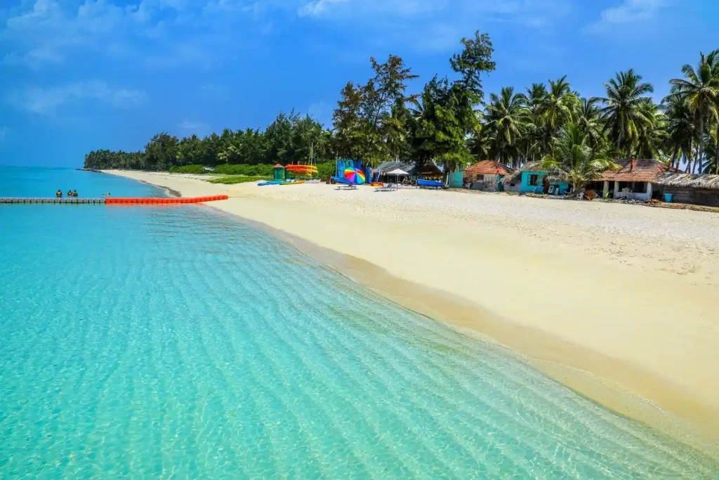 A pristine beach with clear turquoise water gently lapping against a wide, sandy shore. In the background, palm trees and small huts line the beach, with some colorful umbrellas adding pops of color under a bright blue sky. A couple of people are visible in the water.