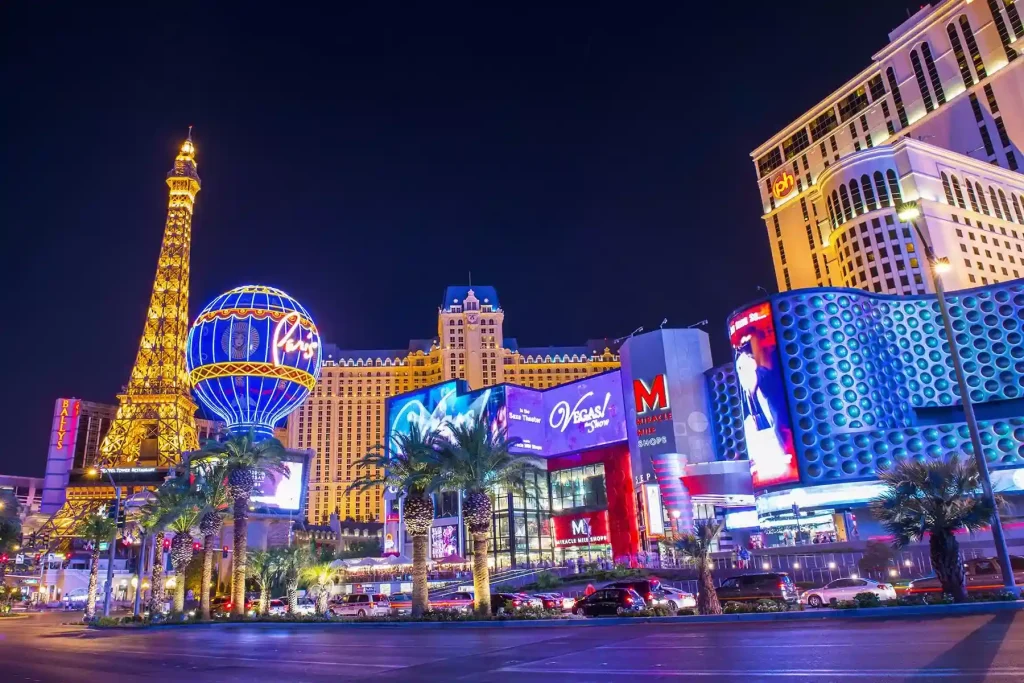 Night view of the Las Vegas Strip, featuring bright neon lights and prominent hotel-casinos. The Eiffel Tower replica at Paris Las Vegas shines beside vibrant signs, including the Planet Hollywood and MGM Grand marquees. Palm trees line the bustling street, epitomizing the essence of Las Vegas.