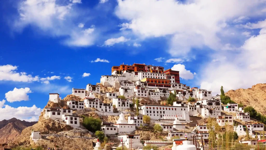 A hilltop monastery with white buildings and red roofs is surrounded by scattered smaller homes against a backdrop of a bright blue sky with fluffy clouds. The structure stands prominently on a rocky hill, showcasing intricate architecture and traditional Buddhist design.