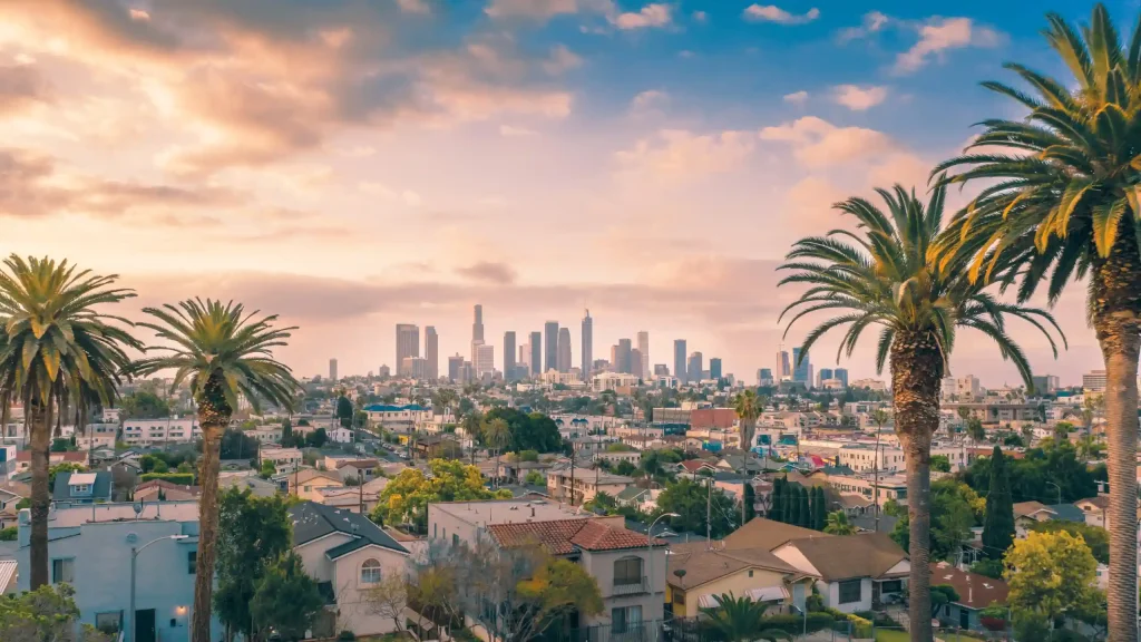 Skyline of Los Angeles with tall buildings in the distance, framed by palm trees. The foreground features residential houses with red-tiled roofs, and the sky above is a mix of soft clouds and pastel hues from the setting or rising California sun.