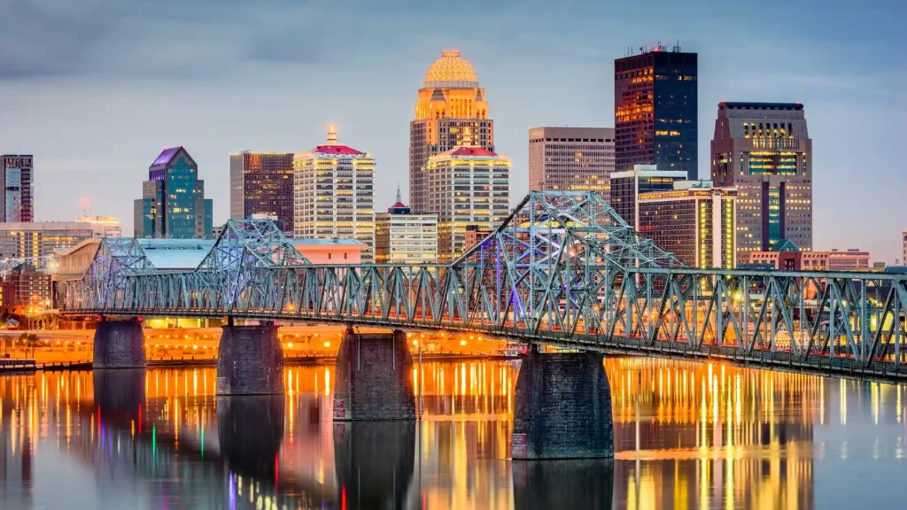 A city skyline at dusk featuring Louisville's prominent, illuminated bridge in the foreground with bright lights reflecting on the water below. Skyscrapers and buildings of various heights, some with colorful lighting, dominate the background under a cloudy evening sky.