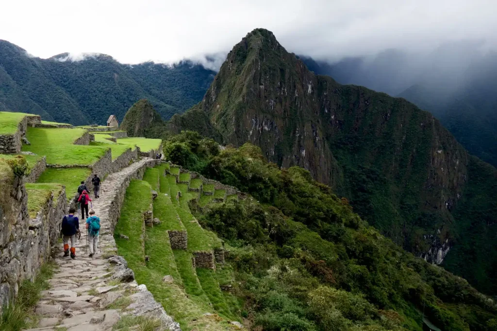 A group of people hike along a stone path surrounded by lush green terraces, reminiscent of Machu Picchu Hiking. In the background, a towering, jagged peak rises into a foggy, cloud-covered sky. Dense forested hills form a dramatic backdrop, creating a serene and adventurous scene