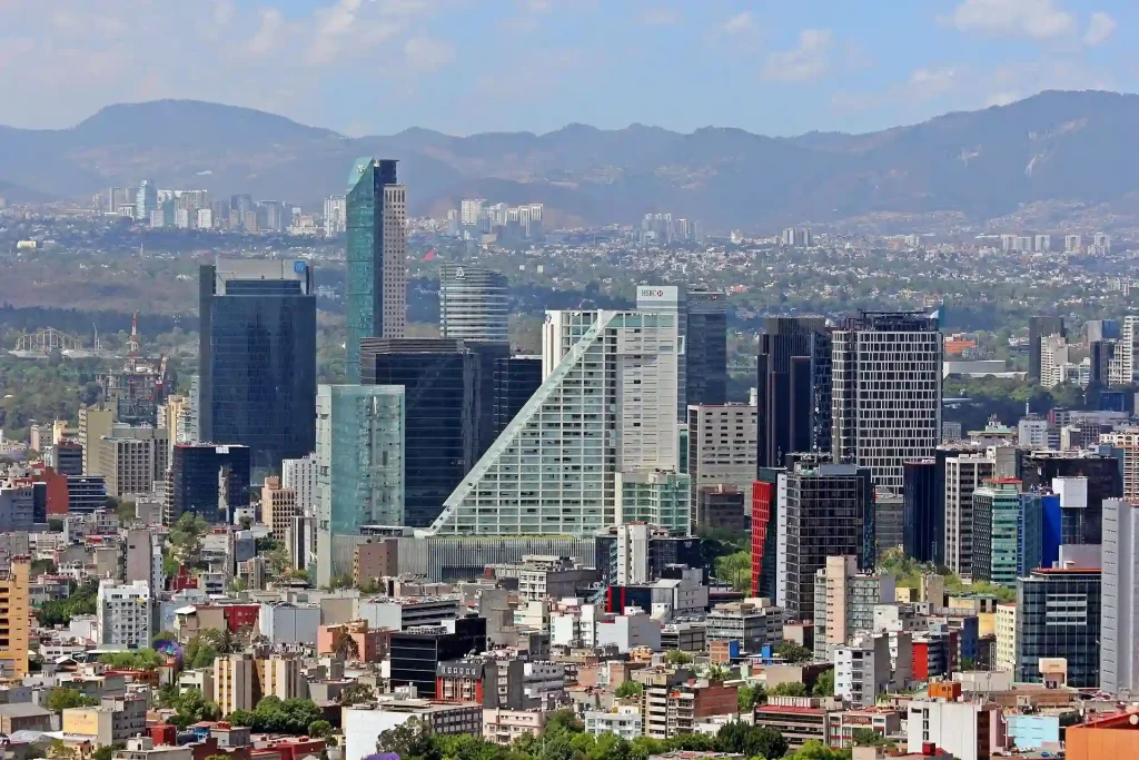 Aerial view of Mexico City's skyline with various high-rise buildings and skyscrapers in the foreground. The landscape includes modern glass buildings, residential areas, and lush greenery. Mountains and a partly cloudy sky are visible in the background.