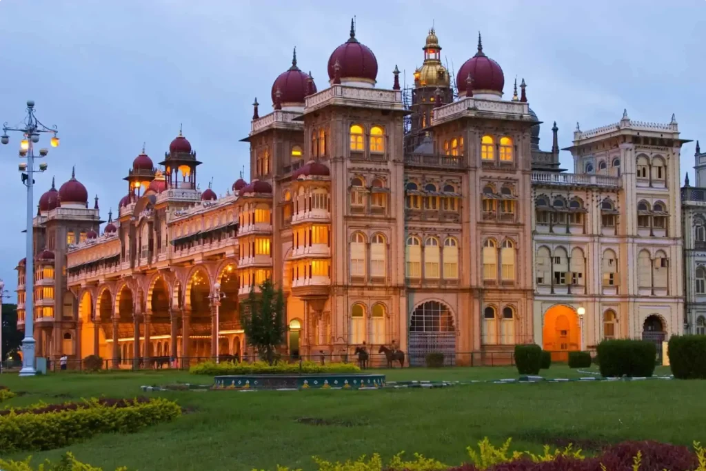 The image shows a grand palace illuminated with warm lights at dusk. The palace features several domes with red tops, intricate architecture, and arched windows. The foreground displays a manicured lawn with hedges and garden paths, creating an enchanting atmosphere.