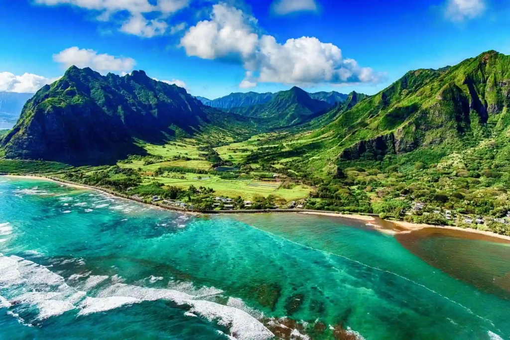 A stunning aerial view of a lush, green valley in Oahu, Hawaii, surrounded by dramatic mountain ranges. The clear, turquoise ocean meets the shore, with white waves breaking along the sandy beach. The landscape is vibrant under a bright blue sky dotted with fluffy white clouds.