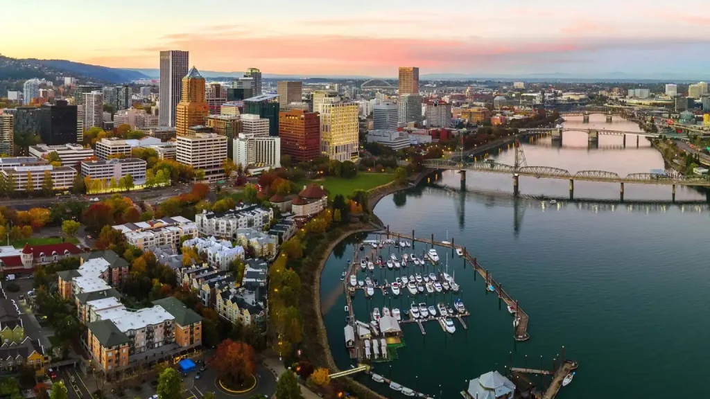 Aerial view of Portland Oregon at sunset featuring a mix of tall buildings and greenery. Below, a waterfront marina with numerous docked boats is visible. To the right, a river runs through the city, crossed by multiple bridges. The sky is painted with soft pastel hues.