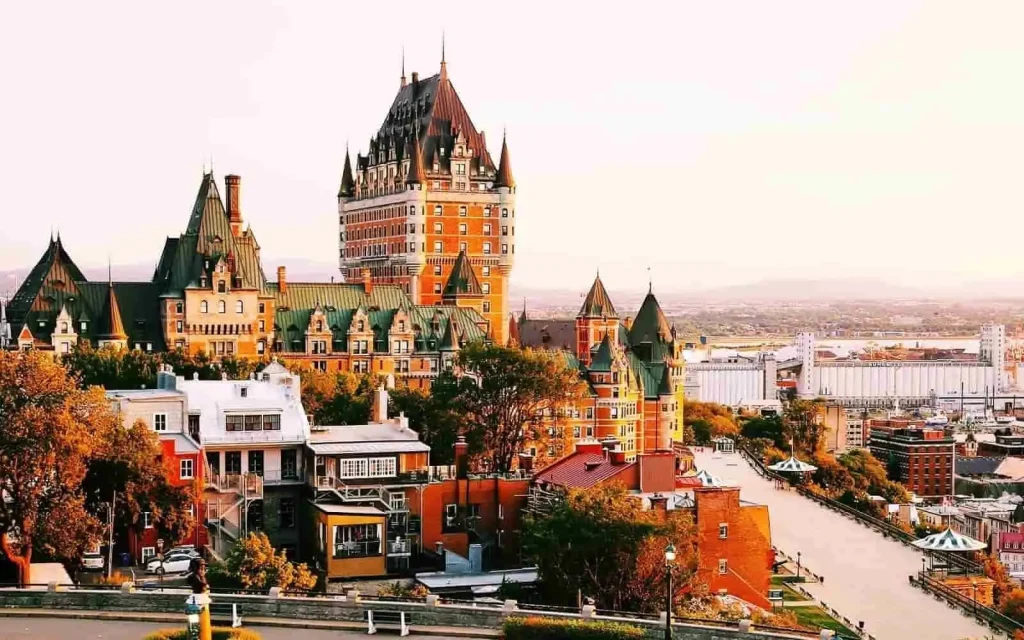 The image shows the Fairmont Le Château Frontenac, a grand hotel with a dramatic, castle-like structure featuring green roofs and a central tower in Quebec City, Canada. The foreground features trees and surrounding buildings, while the background reveals a scenic cityscape under a warm sky.