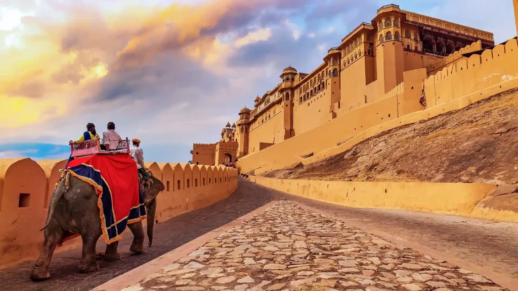 A scenic view of the Amber Fort in Jaipur, India, as seen from a cobblestone path leading up to the fort. An elephant adorned with vibrant red cloth carries two riders towards the entrance. The sky is partly cloudy, creating a picturesque backdrop.