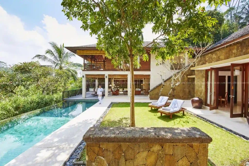 A tropical villa with a Santa Fe touch features a spacious yard with sun loungers, a large swimming pool, and lush green surroundings. The villa has two stories, an open-plan design on the ground floor, and a balcony on the upper level. Trees and plants provide a serene atmosphere.