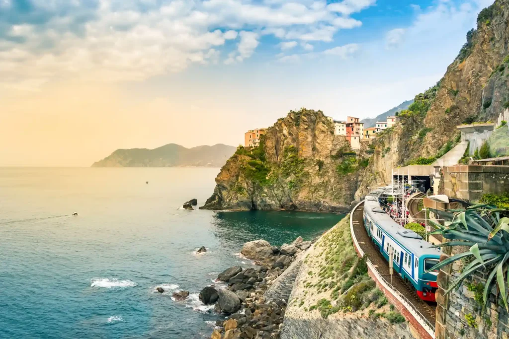 A scenic coastal train travels along a rocky cliffside with the turquoise sea on the left. Colorful buildings are perched on the cliff with mountains in the background under a partly cloudy sky. This picturesque scene showcases the beauty of Italy's coastline as the train moves towards the horizon.