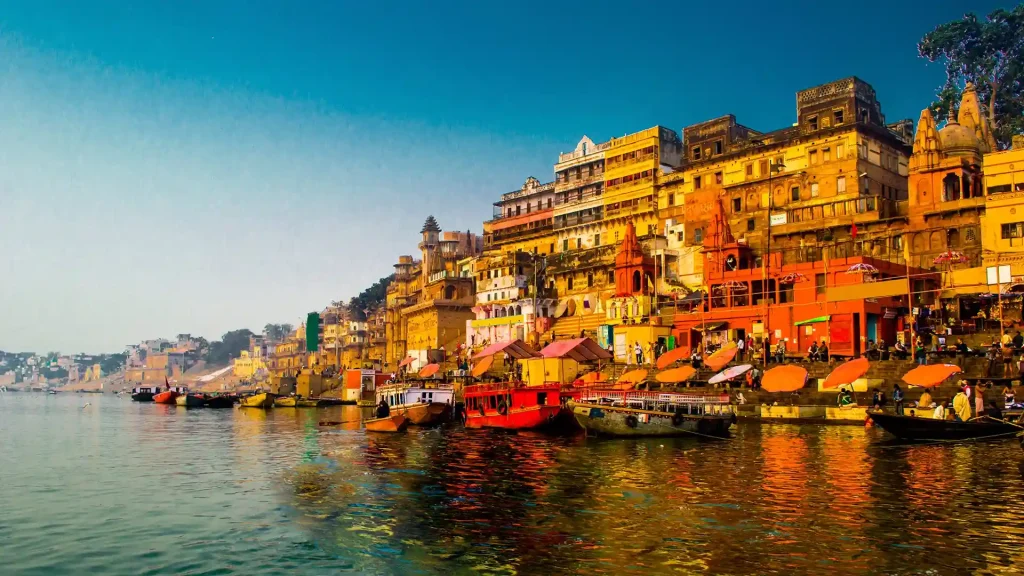 A vibrant riverside scene in India features an array of colorful boats docked along the Ganges River. The shore is lined with historic buildings and ghats, bustling with activity against a clear blue sky. The water mirrors the vivid colors of the surrounding structures, creating a picturesque view.