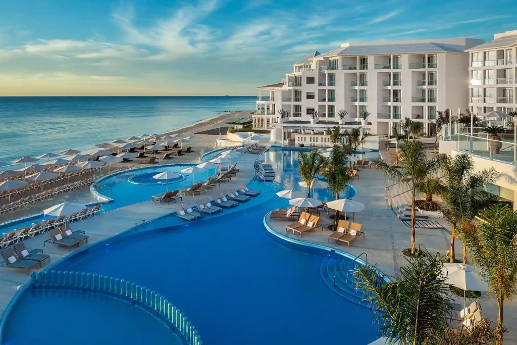 A luxurious beachfront visit to an all-inclusive resort featuring a large, curvy swimming pool surrounded by lounge chairs and palm trees. The modern, multi-story hotel building overlooks the ocean, and the sky is filled with light clouds during sunset.