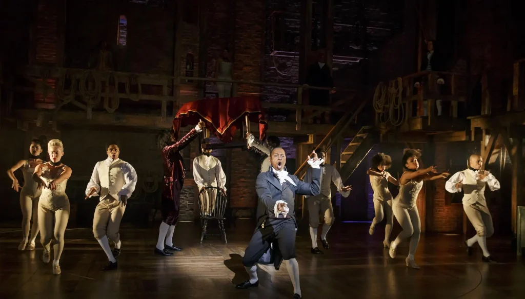 A group of performers dances energetically on a wooden stage with a rustic set behind them, reminiscent of colonial architecture. Watch a Broadway performance as one central performer sings passionately while others move dynamically around him, with a red curtain being raised in the background.