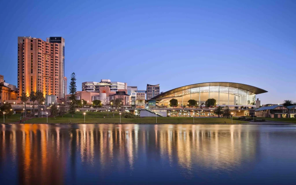 Skyline at dusk with illuminated buildings reflecting in a calm body of water. A prominent glass-domed structure is visible on the right, while several taller buildings stand to the left. The sky is clear with a gradient of blue shades, reminiscent of Adelaide's serene evening vistas.