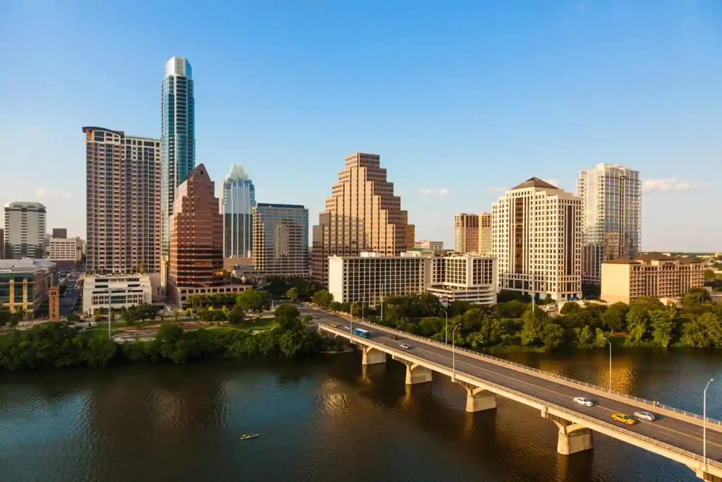 A scenic cityscape view of downtown Austin, Texas, featuring numerous high-rise buildings under a clear blue sky. The city's skyline includes a mix of modern and distinctive architectural styles. In the foreground, cars travel across a bridge over the calm river in Texas.