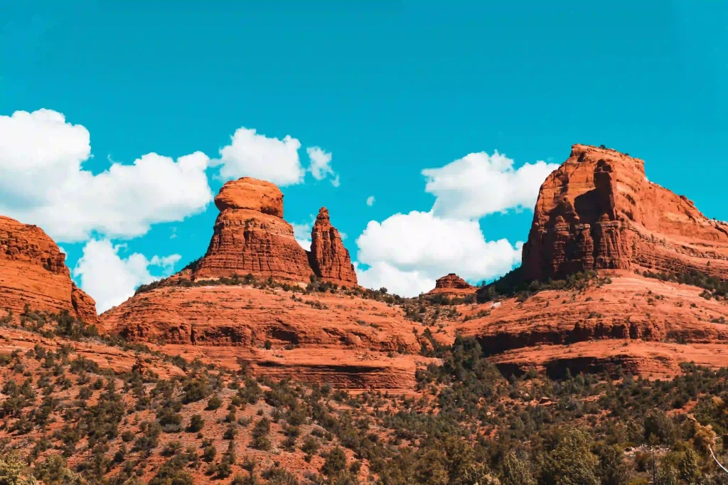 A picturesque view of Sedona's red rock formations under a bright blue sky dotted with fluffy white clouds greets hikers on the Big Park Loop. The rugged terrain, featuring prominent rock structures and scattered green vegetation, creates a stunning contrast against the clear daytime sky.