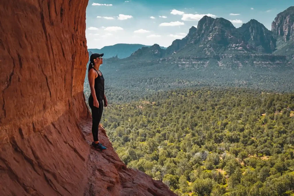 A person in athletic wear stands on a rocky ledge, looking out over a vast, forested valley with a backdrop of rugged mountains beneath a partly cloudy sky. The scene captures a sense of adventure and natural beauty near the renowned Birthing Cave.