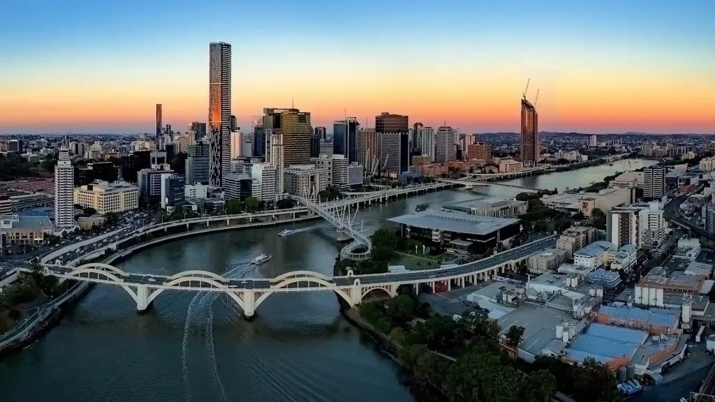 A panoramic view of the Brisbane city skyline at sunset, with tall buildings reflecting in the river below. The scene includes several bridges crossing the river, boats cruising on the water, and a gradient sky with a blend of orange and blue hues transitioning to dusk.