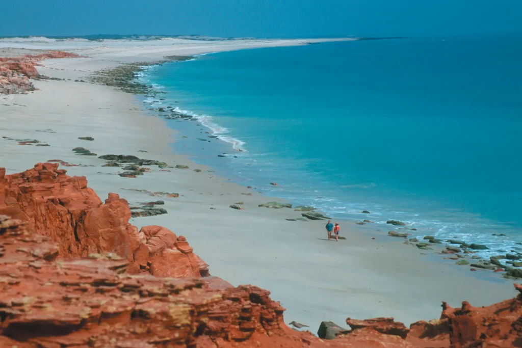 A vast, serene beach with calm turquoise waters and red rocky cliffs in Broome. Two individuals walk along the sandy shore, accompanied by scattered rocks and gently lapping waves. The sky is clear, creating a tranquil, picturesque coastal scene.