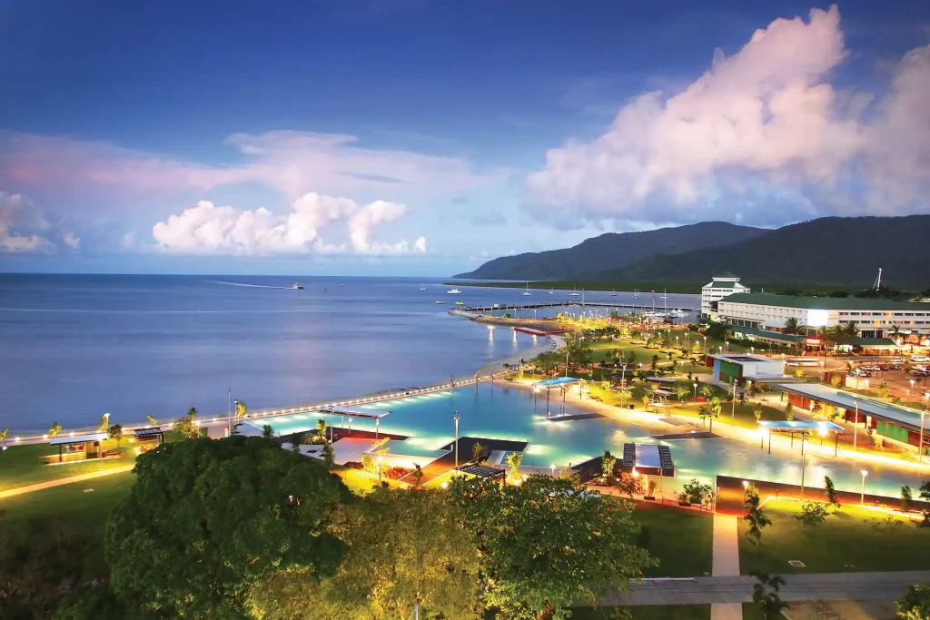 A picturesque seaside view during twilight in Cairns features a tranquil bay, vibrant waterfront, illuminated buildings, and a large pool surrounded by lush greenery. Hills rise in the background under a partly cloudy sky.