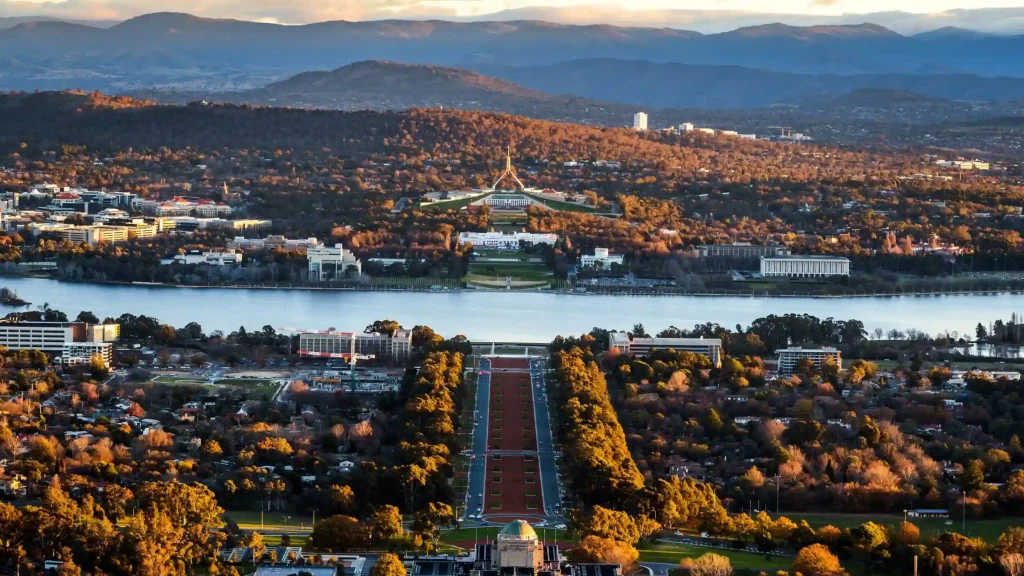 Aerial view of Canberra during sunset, highlighting the central alignment of Anzac Parade leading to the Australian War Memorial in the foreground, with Lake Burley Griffin in the middle and Parliament House set against a backdrop of mountains on the horizon.