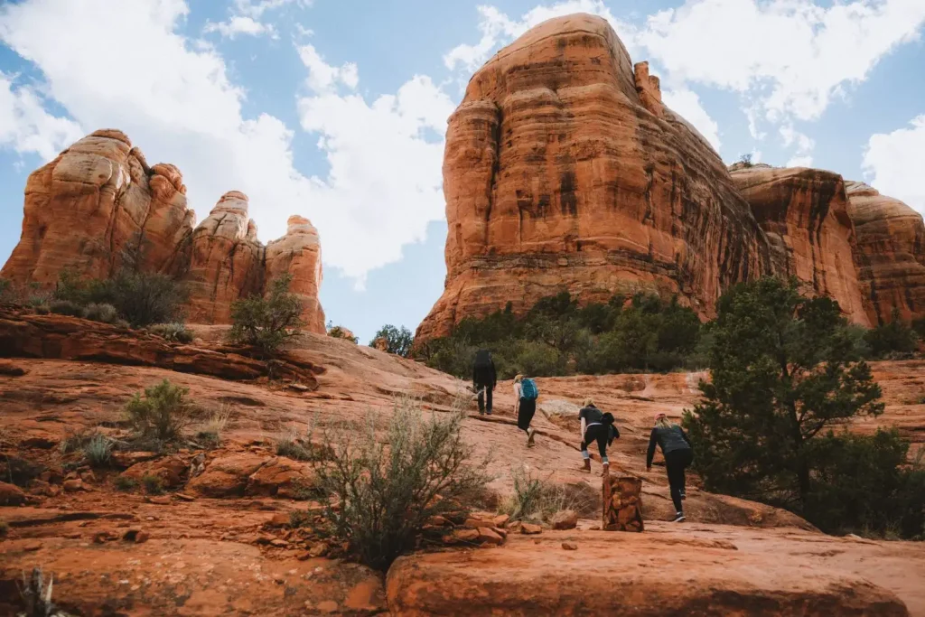 A group of people hiking up the rocky, red sandstone Cathedral Rock Trail, surrounded by towering rock formations and sparse vegetation. The sky is partly cloudy.