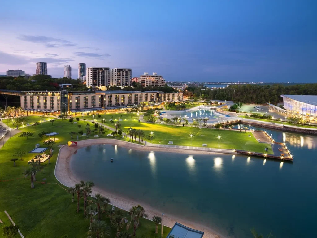 A bird's-eye view of Darwin at dusk reveals a coastal cityscape featuring illuminated modern buildings, a green park with pathways and fountains, and a sandy beach next to calm water. The sky transitions from twilight to night, adding a serene ambiance to the scene.