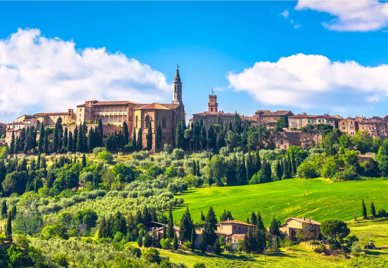 A picturesque hillside town is adorned with age-old buildings and a prominent church with a tall steeple. Lush green fields and trees spread in the foreground under a bright blue sky dotted with fluffy white clouds. Sunny weather enhances the vibrant colors.