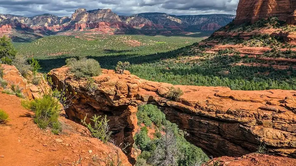 The image shows an expansive view along the Devil's Bridge Trail in Sedona, Arizona. The large sandstone arch stretches across a deep ravine with vibrant red and green vegetation below. Rugged cliffs and forested areas extend into the distance under a partly cloudy sky.