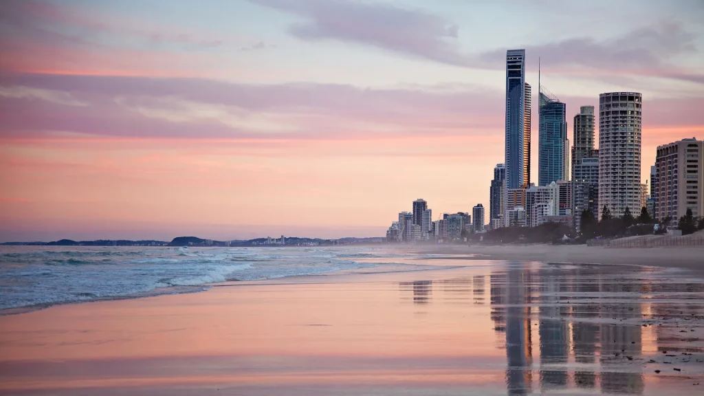 A beach at sunset with gentle waves washing ashore. In the background, several tall skyscrapers and buildings line the Gold Coast under a pink and purple sky. The wet sand reflects the sky and buildings, creating a serene and picturesque scene.