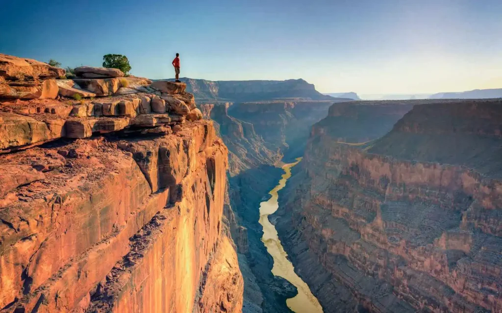 A person stands at the edge of a high cliff, gazing over the vast, winding Grand Canyon below. The scene features steep, rocky walls and a river flowing through the bottom of the canyon in Arizona, with a bright, clear sky above.