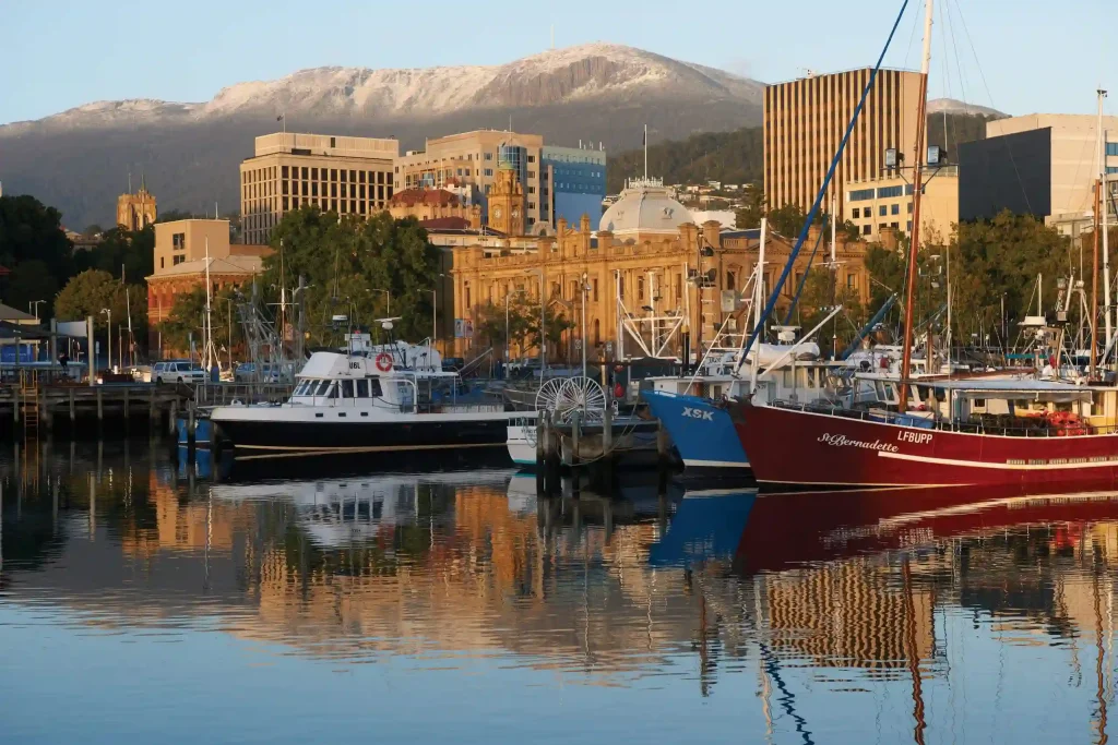 A picturesque view of Hobart's waterfront features several boats docked at a calm harbor. Behind the waterfront, historic and modern buildings stand against a mountainous backdrop with snow-capped peaks under a clear sky.