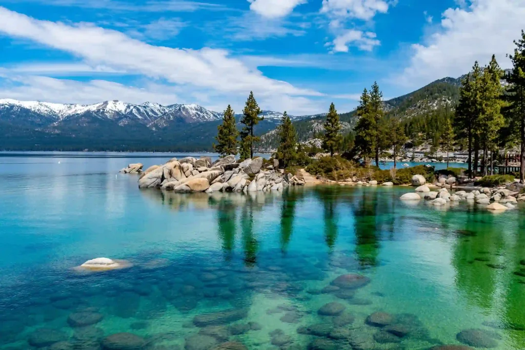 Lake Tahoe boasts a serene expanse of crystal-clear turquoise water with visible rocks beneath the surface. Towering pine trees stand on rocky outcrops by the lakeside, while snow-capped mountains rise majestically in the background under a partly cloudy Nevada sky.