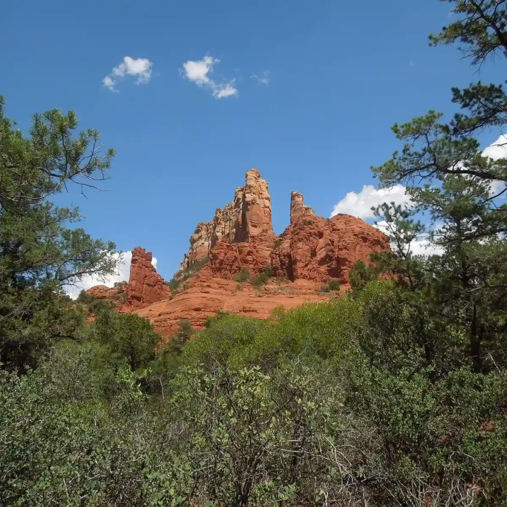 Rock formations in Sedona, Arizona, rise against a blue sky with a few clouds. The rugged, red-hued cliffs are surrounded by green trees and shrubs along Margs Draw Trail, creating a vivid contrast. The natural landscape exudes a sense of tranquility and beauty.