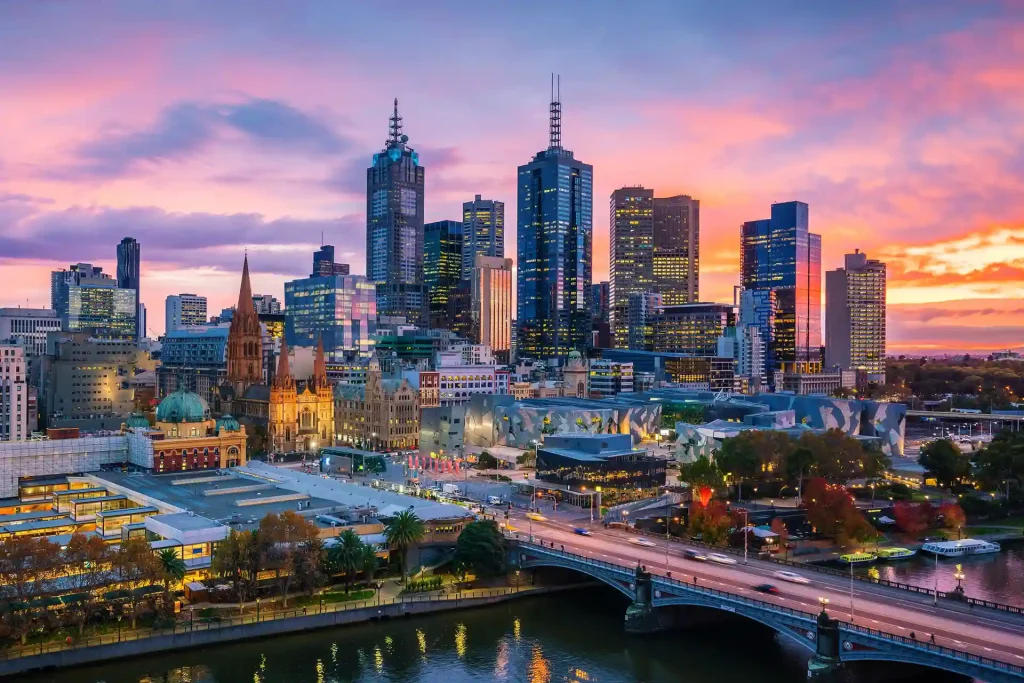 A vibrant cityscape of Melbourne at sunset. Tall skyscrapers dominate the skyline with a mix of modern and historic buildings. A bridge spans across the Yarra River, and colorful lights from buildings reflect in the water as the sky glows with pink and purple hues.