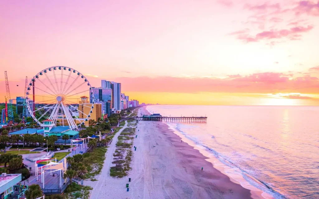 A coastal cityscape at sunset in Myrtle Beach, South Carolina, with a Ferris wheel, tall buildings, and a pier extending into the ocean. The sky is pink and orange, and the beach is mostly empty with a few people strolling along the shore.