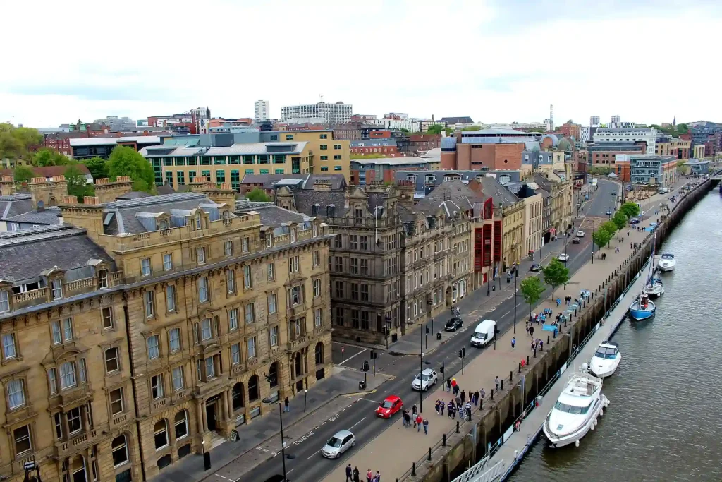 Aerial view of Newcastle's Quayside showing historic and modern buildings lined along the River Tyne. A promenade with people walking and several parked cars can be seen, with boats docked on the river and a cloudy sky in the background, capturing the essence of Newcastle's vibrant waterfront.