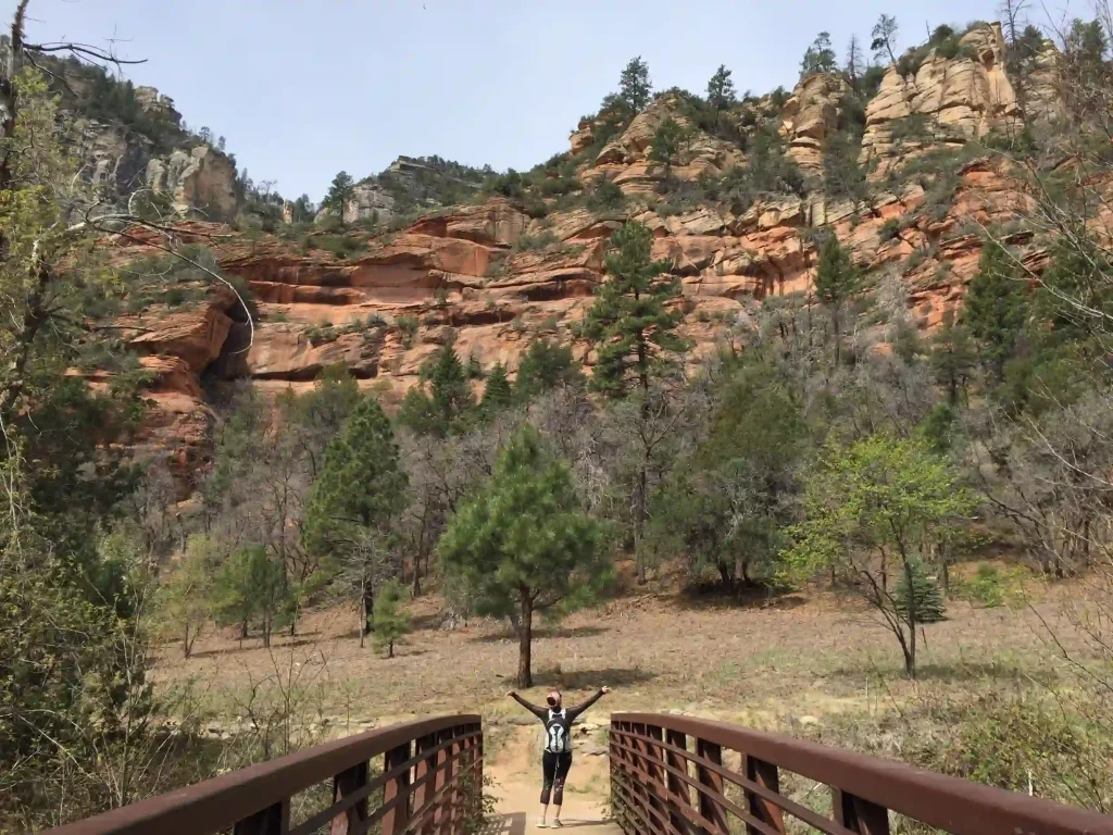 A person stands at the end of a wooden bridge on Oak Creek Trail with arms raised, facing a rocky hillside with patches of greenery. The landscape features red and orange rock formations, sparse trees, and a cloudy sky.